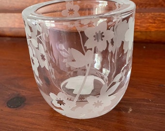 May include: A clear glass votive candle holder with etched floral designs. The glass is curved and features a series of flowers and leaves around the exterior. The candle holder sits on a wooden surface.