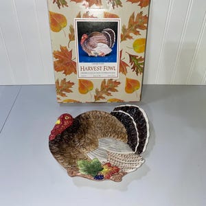 May include: A ceramic turkey-shaped serving plate with a brown, white, and black design. The plate has a red head and a fan-shaped tail. The box behind the plate has a fall leaf design and the words "HARVEST FOWL".