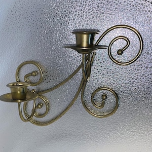 May include: A brass wall sconce with two candle holders and decorative scrollwork. The sconce has a vintage aesthetic, with a warm, golden hue. The design features elegant curves and small ball accents, suitable for home decor.