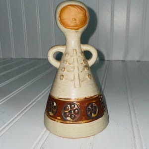 May include: A vintage ceramic figurine in a cream color with brown accents. The figurine has a rounded head with a face, two handles, and a conical body. The body features a brown band with floral designs and a vertical tree-like pattern.
