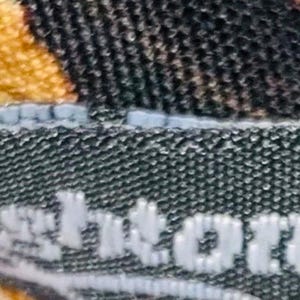 Pode incluir: Close-up de uma etiqueta de tecido preta com a palavra "Brighton" em letras brancas. A etiqueta &eacute; costurada em um tecido com um padr&atilde;o de estampa de animal marrom e amarelo.
