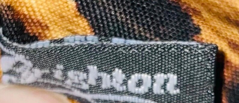 Pode incluir: Close-up de uma etiqueta de tecido preta com a palavra "Brighton" em letras brancas. A etiqueta &eacute; costurada em um tecido com um padr&atilde;o de estampa de animal marrom e amarelo.
