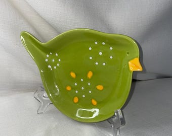 May include: A green ceramic bird-shaped dish with white and yellow polka dots. The dish is on a clear plastic stand.