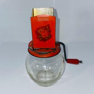 May include: Vintage glass jar with a red and gold metal grinder. The grinder has a red handle and a red metal top with a floral design. The jar is clear and empty.