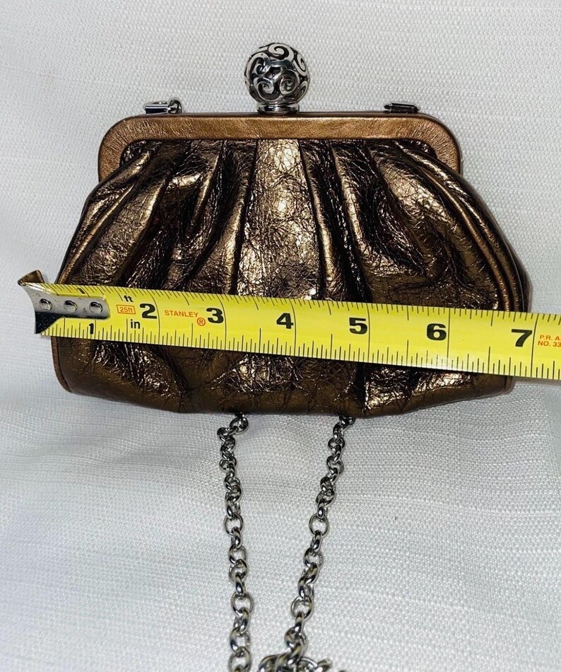 Pode incluir: Uma clutch de cor bronze com uma al&ccedil;a de corrente prateada. A clutch tem aproximadamente 18 cm de largura. Possui um fecho de bola prateada decorativo na parte superior. A bolsa tem uma apar&ecirc;ncia texturizada e enrugada.