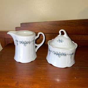 May include: A white creamer and sugar bowl set with a blue floral pattern. The creamer has a handle and a spout. The sugar bowl has a lid.