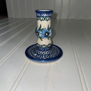 May include: A ceramic candle holder with a blue and white floral design. The base is a small, round plate with a detailed pattern. The body of the holder is cylindrical, with a bulbous middle section and a flared top. The color scheme is primarily blue and white.