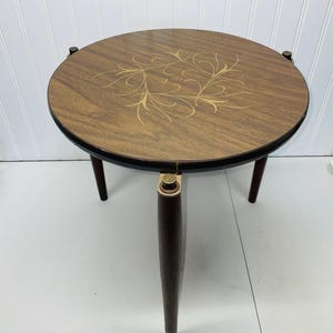 May include: A round side table with a wood grain top featuring a gold-colored swirling leaf pattern. The table has three dark brown tapered legs attached to the circular frame with gold-colored metal hardware accents at the top of each leg.