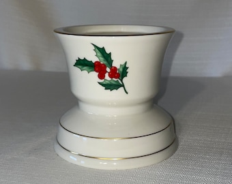 May include: A white ceramic candle holder with a holly and berry design. The candle holder has a flared top and a round base, with gold trim around the top and base. The holly leaves are green, and the berries are red.