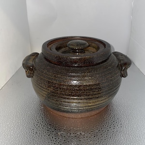 May include: A brown ceramic pot with a lid. The pot has two handles on the sides and a knob on the lid. The pot is approximately 8 inches in diameter and 6 inches tall.