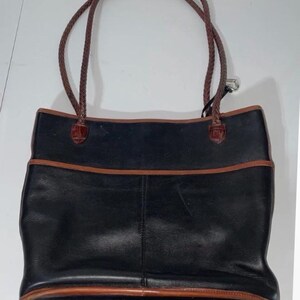 May include: Black leather tote bag with brown trim and braided brown handles. The bag has a rectangular shape with a pocket on the front. The handles are attached with brown leather accents.