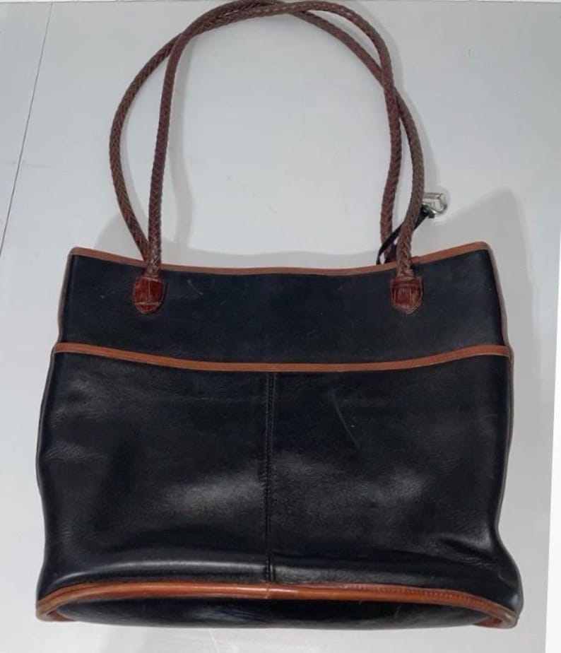 May include: Black leather tote bag with brown trim and braided brown handles. The bag has a rectangular shape with a pocket on the front. The handles are attached with brown leather accents.