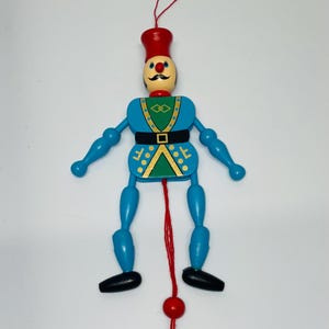 May include: A wooden jumping jack toy featuring a soldier figure with a red hat, blue limbs, and a blue and green torso. The figure has a painted face with a black mustache and a red nose. A red string hangs from the bottom with a red bead.