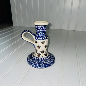 May include: A ceramic candle holder with a handle, featuring a blue and white polka dot pattern. The base and handle are adorned with a blue and white design, while the body has a pattern of brown fruit with green leaves.