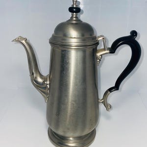May include: A vintage silver-toned coffee pot with a black handle and lid finial. The pot features a curved spout and ornate detailing on the handle attachments. The design is reminiscent of antique kitchenware.