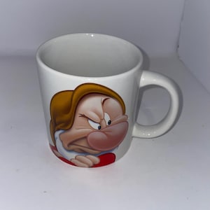 May include: White ceramic mug featuring a cartoon image of Grumpy from Snow White. The mug has a curved handle and a red heart-shaped detail on the bottom of the character's clothing. The character has a grumpy expression.