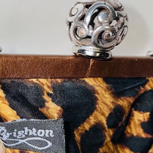 Pode incluir: Um fecho esf&eacute;rico vazado prateado fica em cima de uma moldura de couro marrom. O tecido interior apresenta uma estampa de leopardo preta e marrom. A etiqueta diz "Brighton".