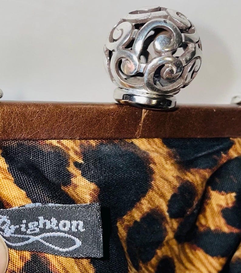 Pode incluir: Um fecho esf&eacute;rico vazado prateado fica em cima de uma moldura de couro marrom. O tecido interior apresenta uma estampa de leopardo preta e marrom. A etiqueta diz "Brighton".