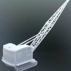 Könnte beinhalten: Graues Modell eines Raupenkrans mit detaillierter Kabine, Gitterausleger und Kettenlaufwerk. Der Ausleger des Krans erstreckt sich in einem Winkel nach oben und zeigt sein kompliziertes Design. Das Modell steht vor einem dunklen Hintergrund.