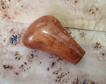 May include: A polished, wooden gear shift knob with a rounded top and tapered base. The knob has a warm, reddish-brown hue with visible wood grain and a glossy finish. The background is a light-colored wood surface.