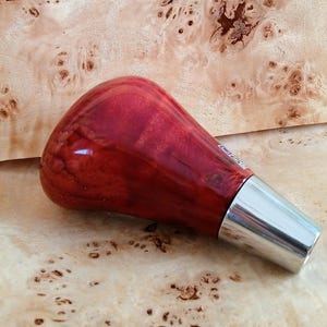 May include: A red wood gear knob with a silver metal base. The knob is made of wood with a red and white marbled pattern.