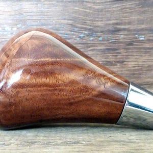 以下が含まれることがあります： 銀色の金属製のトリムが付いた茶色の木製シフトノブ。ノブは滑らかで磨かれた仕上げで、独特の木目模様があります。