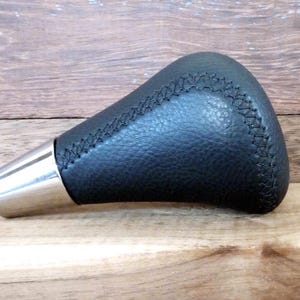 May include: A black gear shift knob featuring a textured leather-like surface with prominent cross-stitching along the edges and a smooth, tapered metallic silver base. The object has a curved, ergonomic shape designed for manual vehicle transmission.