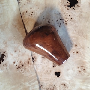 Könnte beinhalten: Ein glänzender, brauner Holzschaltknauf mit abgerundeter Oberseite und konischem Fuß. Der Knauf befindet sich auf einer hellen, gemusterten Holzoberfläche, wodurch ein Kontrast in Texturen und Tönen entsteht.