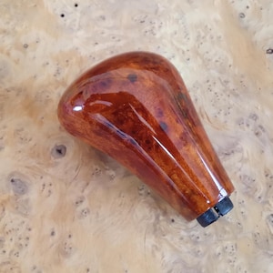 Puede incluir: Una perilla de cambio de marchas de madera marrón con un acabado brillante. La perilla tiene un patrón de veta de madera único y una base de plástico negro.