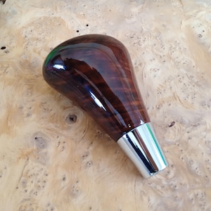 Könnte beinhalten: Ein Schaltknauf mit einer polierten, dunkelbraunen Holzmaserung und einer glatten, glänzenden Oberfläche. Die Basis des Knaufs besteht aus einer konischen, reflektierenden silberfarbenen Metallmanschette, die mit dem oberen Holzteil verbunden ist.