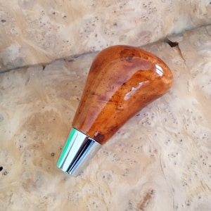 May include: A wooden gear shift knob with a glossy finish. The knob is a warm brown color with a silver metal base. The wood grain is visible, adding texture to the smooth surface. The knob is designed for a vehicle's gear shift.
