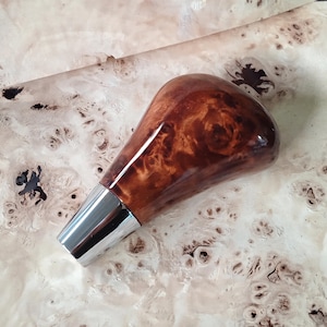 May include: A wooden gear shift knob with a glossy, brown finish and a silver-colored base. The wood grain has a unique pattern. The knob is set against a light-colored, textured background.