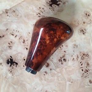 Puede incluir: Un pomo de palanca de cambios de madera brillante con un rico patrón de veta de madera de nogal marrón. El pomo tiene una parte superior redondeada y una forma cónica, con una base de metal oscuro. El fondo es una superficie de madera clara con una textura única y moteada.