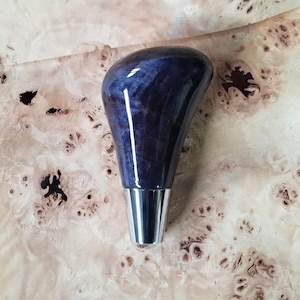May include: A gear shift knob featuring a polished, deep purple, swirling wood grain pattern with a smooth, glossy finish. The top section is bulbous and tapers downward into a sleek, silver-toned metallic base that is cylindrical in shape.
