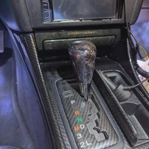 May include: A car's interior showing a gear shift lever with a dark, marbled finish. The shift console has a carbon fibre pattern, with gear positions marked P, R, N, D, 3, and 2L. The dashboard includes a display screen and the text "SPORTDESIGN".