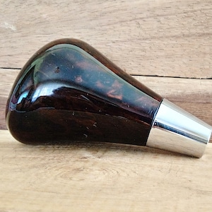May include: A dark brown, teardrop-shaped gear shift knob with a glossy finish. The top portion has a marbled appearance, and the base is silver. The knob is set against a wooden background.
