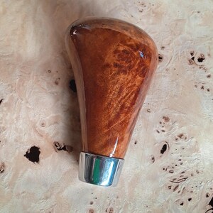 Könnte beinhalten: Ein hölzerner Schaltknauf mit glänzendem Finish. Der Knauf hat eine warme braune Farbe mit einem silbernen Sockel. Die Holzmaserung ist sichtbar und verleiht dem Design Textur. Der Knauf ist wahrscheinlich für ein Auto.