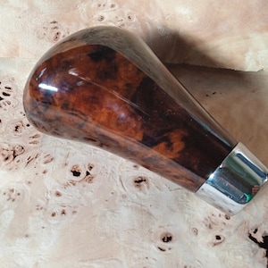 Puede incluir: Un pomo de palanca de cambios de madera en forma de lágrima con un acabado brillante. El pomo es principalmente marrón con patrones de vetas de madera más oscuros y una base de metal plateado. El artículo es probablemente para un vehículo.