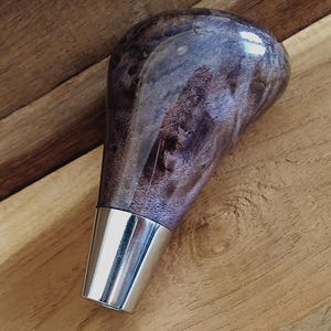 Puede incluir: Un pomo de palanca de cambios artesanal de madera de raíz morada y gris, con un acabado pulido y brillante. El pomo tiene una base de metal plateado. El patrón de la veta de la madera es visible. Diseñado para un vehículo.