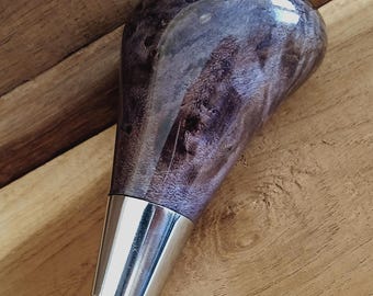 May include: A handcrafted, purple and gray burl wood shift knob with a polished, glossy finish. The knob has a silver-colored metal base. The wood grain pattern is visible. The shift knob is designed for a vehicle.