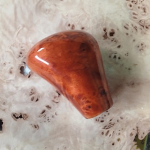 May include: A polished, reddish-brown wooden knob with a glossy finish. The knob has a rounded top and a tapered base, showcasing the natural wood grain. The background is a light-colored surface with a unique, speckled pattern.