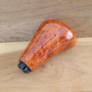 Könnte beinhalten: Ein hölzerner Schalthebelknauf für ein Auto mit glänzendem Finish. Der Knauf hat eine warme, rotbraune Farbe mit einem einzigartigen Holzmaserungsmuster. Die Basis des Knaufs ist schwarz.