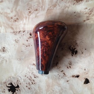 Puede incluir: Un pomo de palanca de cambios de madera brillante con un rico patrón de vetas de madera de color marrón rojizo. El pomo tiene una parte superior redondeada y un cuerpo cónico, con una base oscura con dos pequeños postes metálicos. El fondo es una superficie de madera clara.