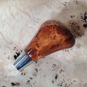 Puede incluir: Un pomo de palanca de cambios de madera pulida con un patrón de veta de madera nudosa en tonos marrones y bronceados, unido a una base de metal cilíndrica de color plateado que se estrecha hacia el extremo inferior del componente.