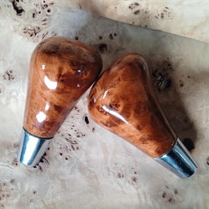 Könnte beinhalten: Zwei Schaltknöpfe aus Holz mit glänzendem Finish. Die Knöpfe haben eine warme braune Farbe mit einem einzigartigen Holzmaserungsmuster. Jeder Knopf hat einen silberfarbenen Metallsockel.