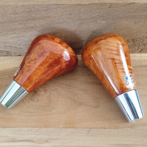 May include: Two gear shift knobs featuring a polished, light brown wood grain finish with a glossy coating. Each knob has a tapered silver metal base and a small square label on the side with the letters C, M, D, and N stacked vertically in blue.