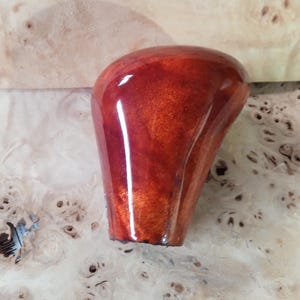 Puede incluir: Un pomo de palanca de cambios de madera, de color marrón rojizo brillante, con una parte superior redondeada y una base cónica. El pomo se encuentra sobre un fondo claro y texturizado, que resalta la veta y la textura natural de la madera.