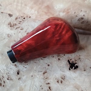 May include: A red burl wood gear knob with a polished finish. The knob has a black base and a smooth, ergonomic shape. The background is a light-coloured wood surface with a unique, natural grain pattern.