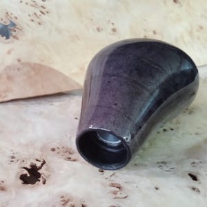 Op de afbeelding: Een donker, glanzend, druppelvormig object met een cilindrische opening aan de basis. Het oppervlak heeft een wervelende, gemarmerde uitstraling in grijstinten en paars. Het object is waarschijnlijk een decoratief of functioneel item.