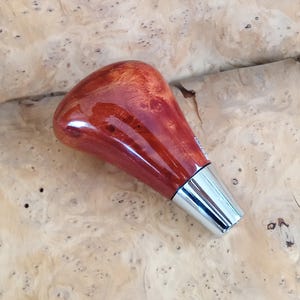 May include: A polished, reddish-brown wooden gear shift knob with a smooth, glossy finish and a prominent burl wood grain pattern. The top section is rounded and bulbous, tapering down to a sleek, silver-coloured metallic base connector.